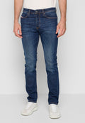Mann in schlank geschnittenen dunkelblauen Jeans und weißen Sneakers, der vor einem schlichten hellen Hintergrund steht.