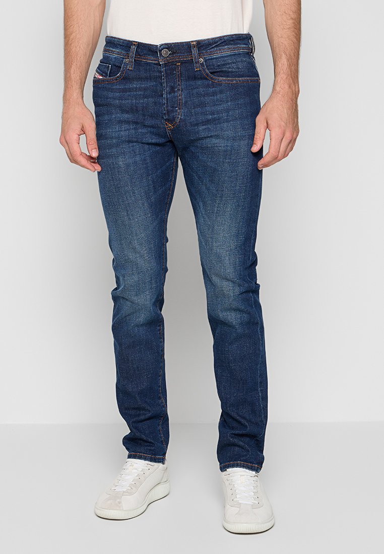 Mann in schlank geschnittenen dunkelblauen Jeans und weißen Sneakers, der vor einem schlichten hellen Hintergrund steht.