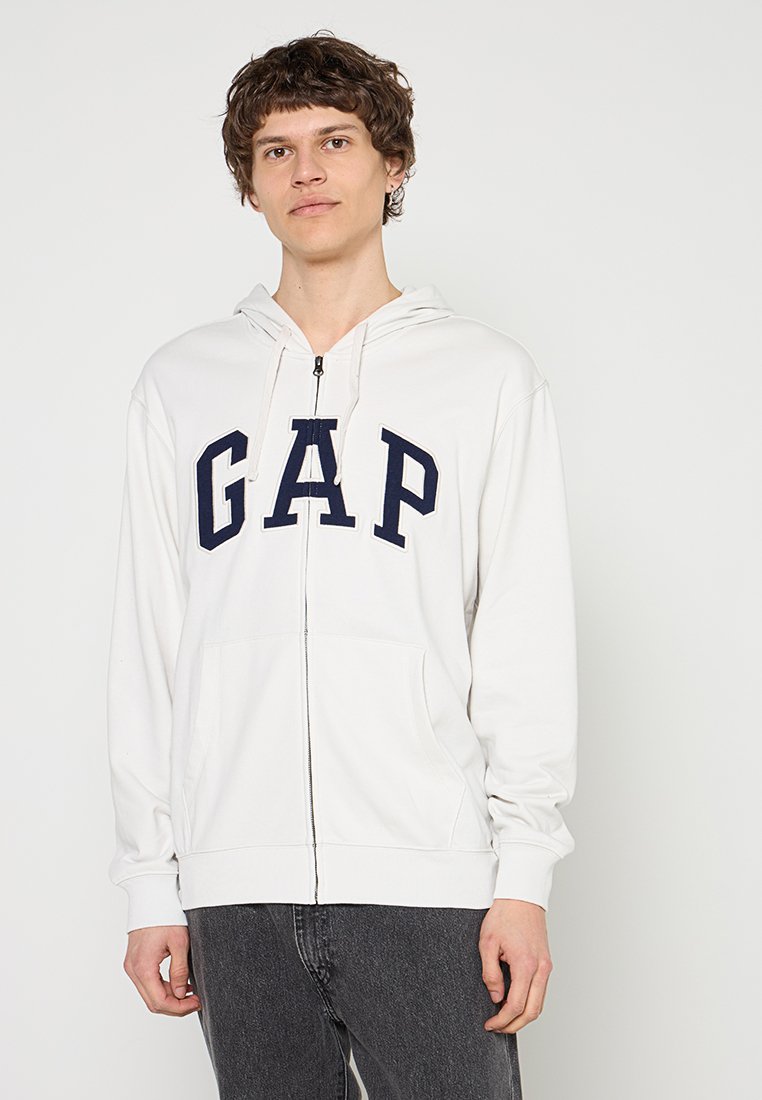 Junger Mann trägt einen weißen Hoodie mit Reißverschluss und dem Schriftzug "GAP" in Marineblau, kombiniert mit dunkelgrauen Jeans, steht vor einem schlichten hellen Hintergrund.