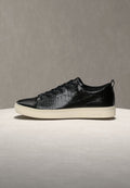 Schwarzer Ledersneaker mit Krokodilstruktur, flacher weißer Sohle, abgerundeter Zehenpartie und schwarzen Schnürsenkeln. Verfügt über eine Zuglasche an der Ferse.