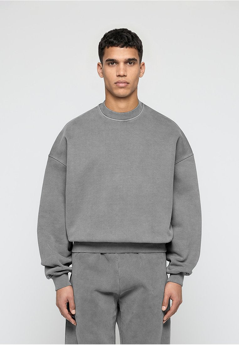 Mann, der nach vorne schaut und einen lockeren grauen Pullover sowie passende graue Hosen vor einem schlichten hellen Hintergrund trägt.