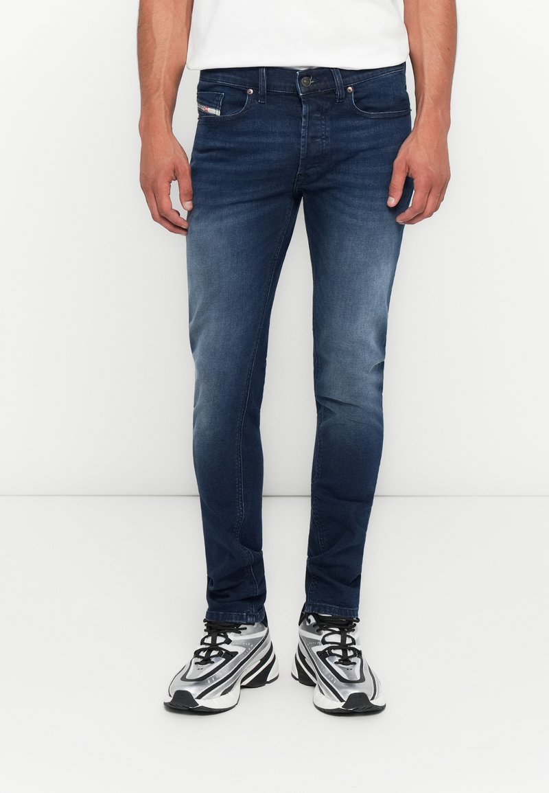 Mann in eng anliegenden dunkelblauen Jeans und schwarz-weißen Sportschuhen, stehend vor einem schlichten weißen Hintergrund.