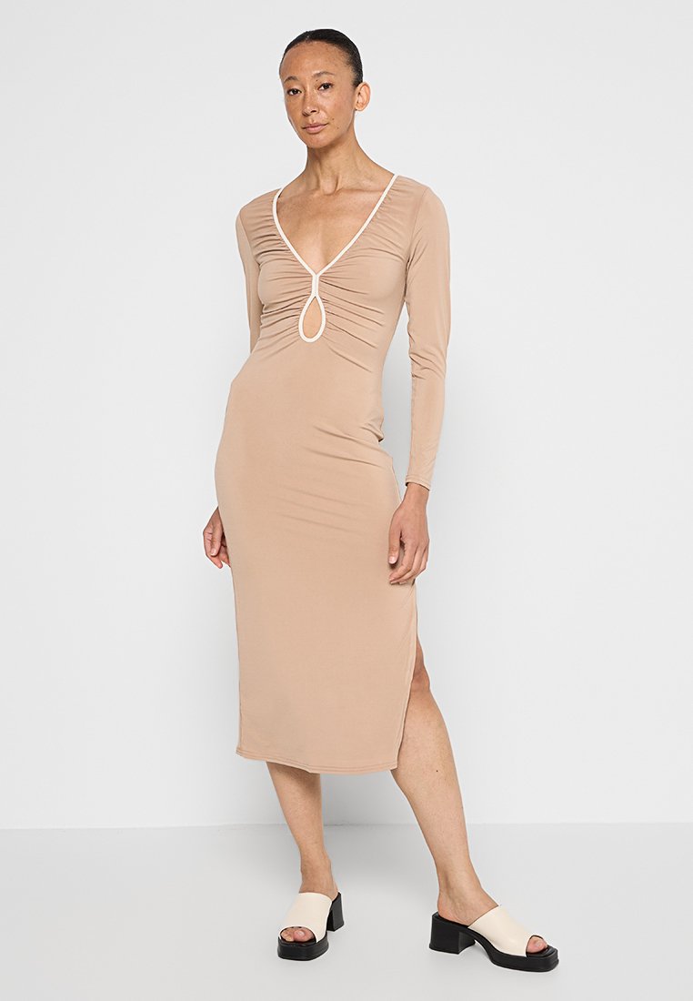 Beiges Langarmkleid mit tiefem V-Ausschnitt und Raffung, ausgestattet mit einem Ausschnittdetail und seitlichem Schlitz. Kombiniert mit schwarzen Plateausandalen.