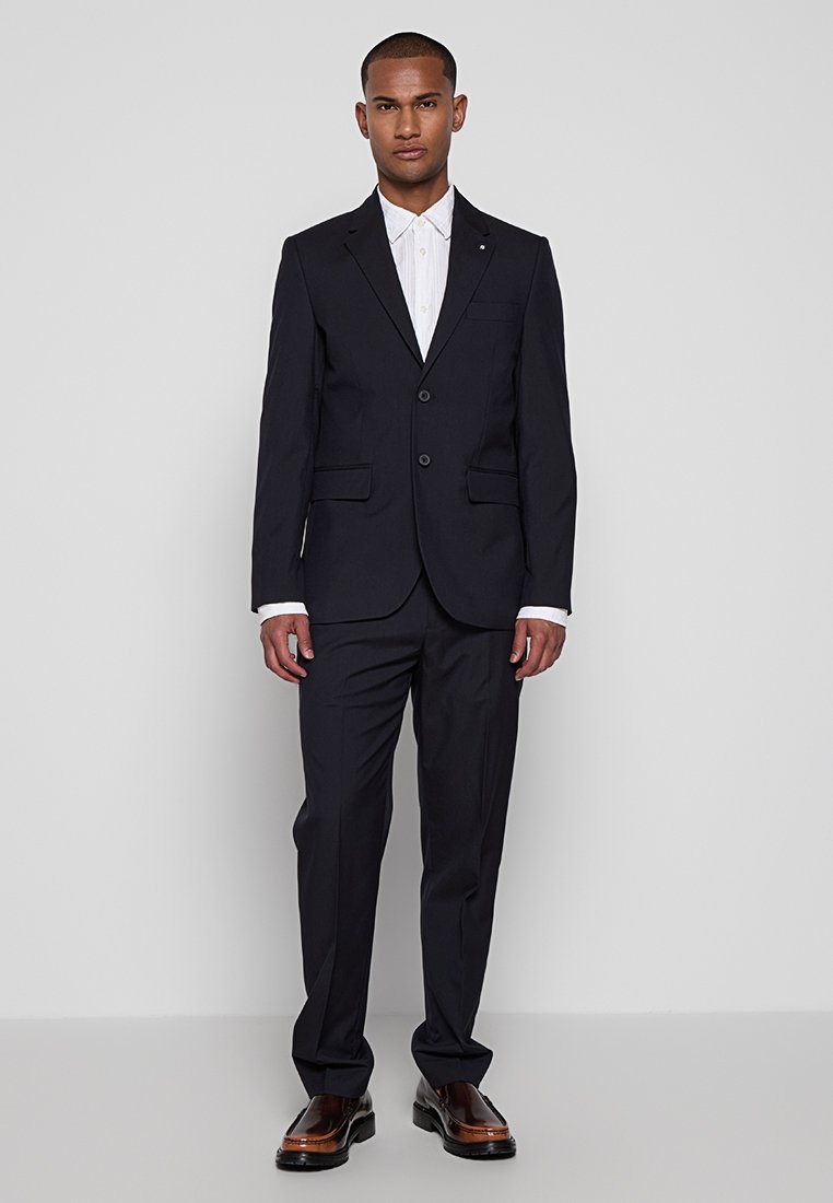 Marineblauer Anzug mit strukturiertem Blazer, zwei Knöpfen und geraden Hosen. Weißes Hemd mit Kragen darunter, lackierte Schuhe.