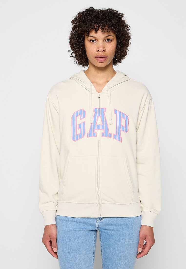 Frau trägt einen cremefarbenen Kapuzenpullover mit Reißverschluss, darauf ein blau-pinkes GAP-Logo, sowie eine hellblaue Jeans und steht vor einem schlichten Hintergrund.