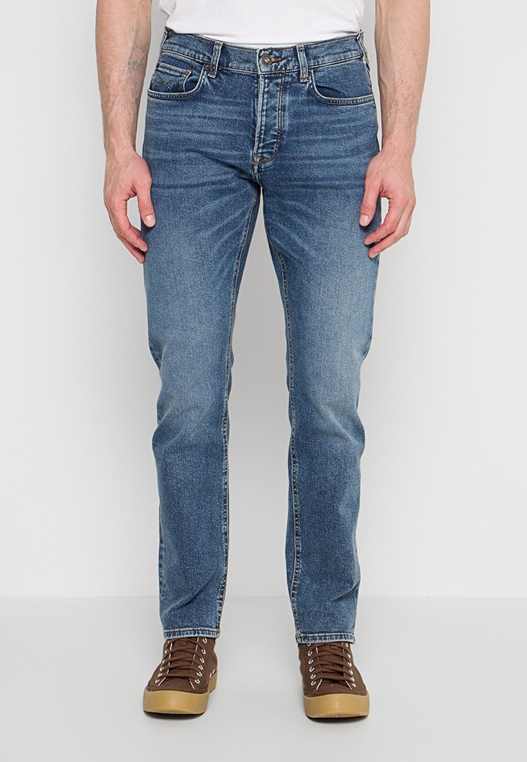 Mann trägt blaue Jeans mit geradem Bein und braune Schnürsneaker, steht vor einem schlichten hellen Hintergrund.