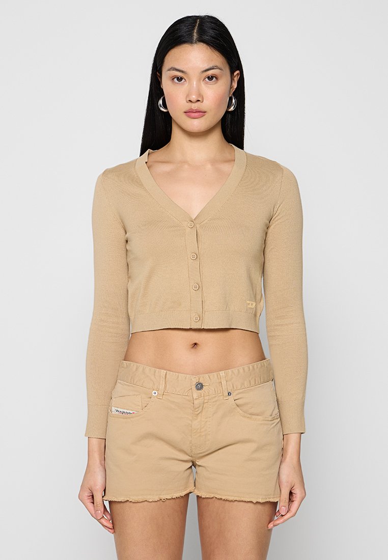 Beige cropped Cardigan mit V-Ausschnitt, langen Ärmeln und Knopfverschluss; kombiniert mit hellbeigen Shorts mit fransigem Saum.