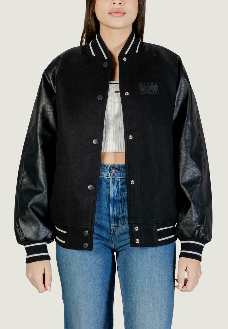Frau trägt eine schwarze College-Jacke mit Ledermanschetten, weißen, kurzen Stricktop und hoch taillierte blaue Jeans, steht vor einem schlichten Hintergrund.