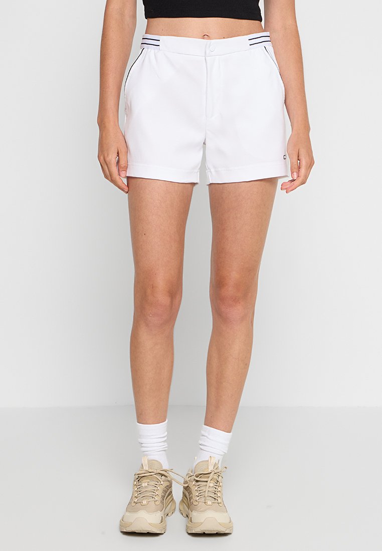 Frau, die weiße Sportshorts mit seitlichen Taschen, beigen Sneakern, weißen Socken und einem schwarzen Cropped-Top trägt, steht vor einem schlichten Hintergrund.