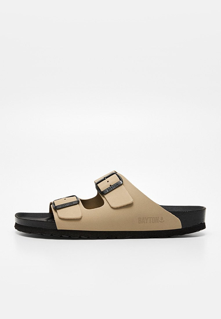 Beige offene Sandale mit zwei verstellbaren schwarzen Metallverschlüssen und schwarzer Sohle, Seitenansicht, mit der Marke "Bayton" auf den Riemen und der Seite.