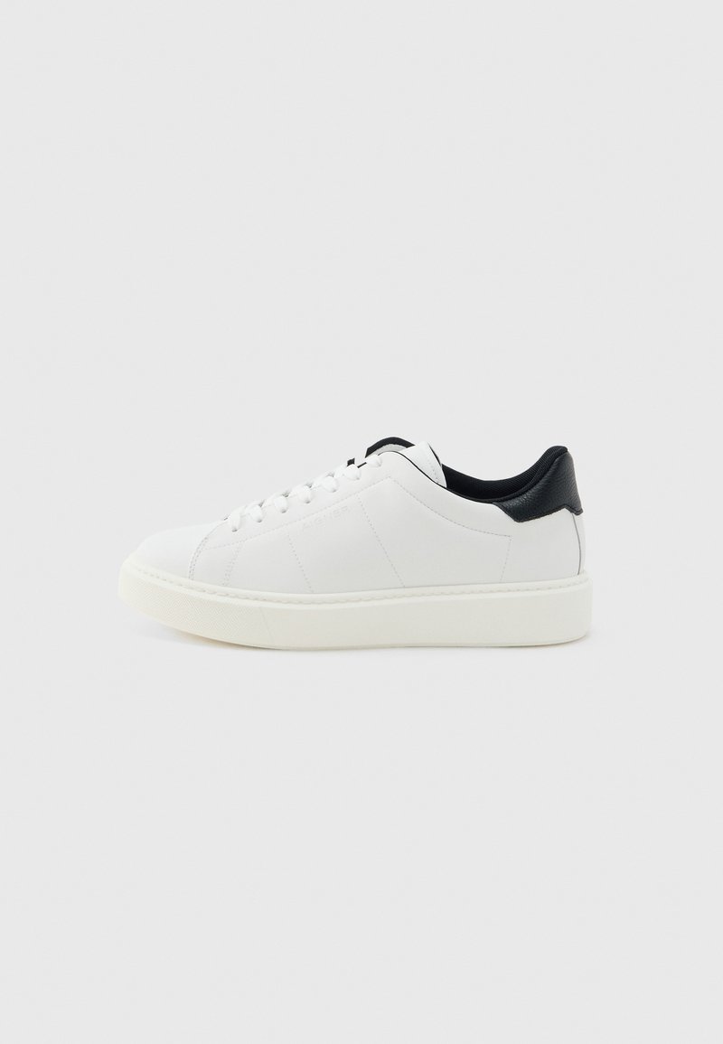 Weißer Sneaker mit niedrigem Schaft, weißen Schnürsenkeln, dicker Sohle und schwarzem Fersenkragen, im Profil vor hellem grauem Hintergrund gezeigt.