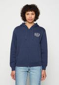 Junge Person mit lockigem Haar, die eine marineblaue GAP-Kapuzenjacke mit Reißverschluss und hellblaue Jeans trägt, vor einem einfarbigen Hintergrund steht.