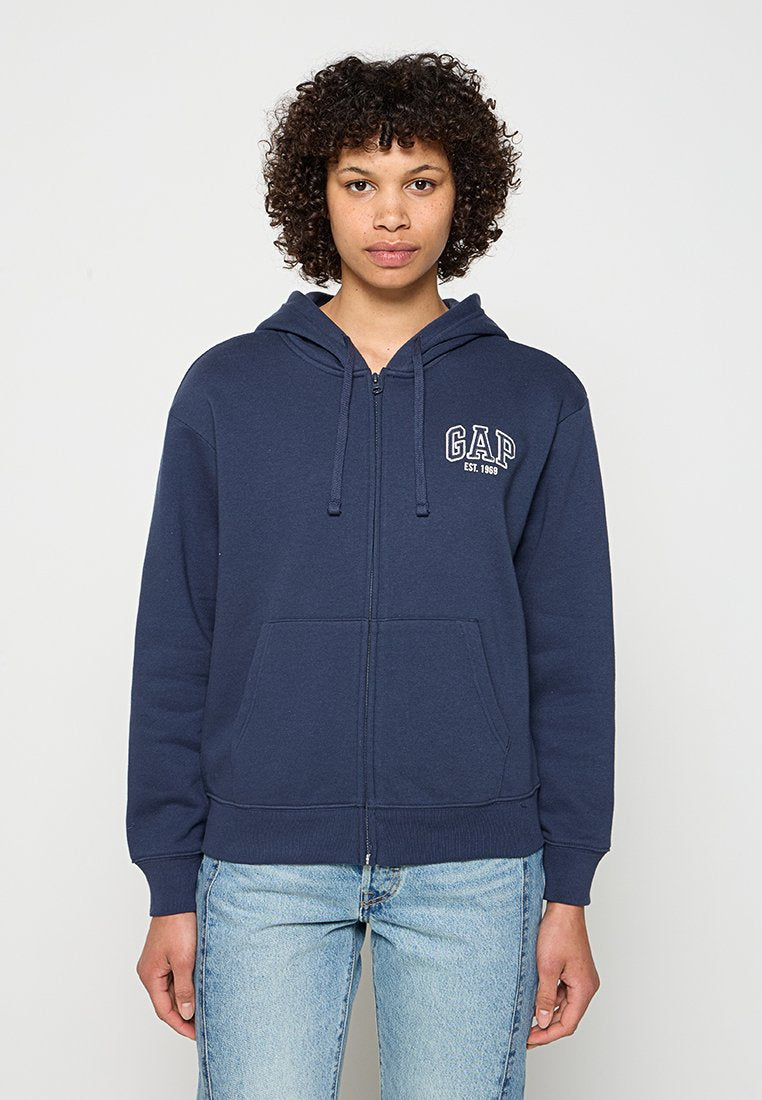 Junge Person mit lockigem Haar, die eine marineblaue GAP-Kapuzenjacke mit Reißverschluss und hellblaue Jeans trägt, vor einem einfarbigen Hintergrund steht.