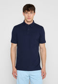 Junger Mann, der ein marineblaues kurzärmeliges Polohemd und hellblaue Hose trägt, steht mit dem Gesicht nach vorne vor einem einfarbigen Hintergrund.