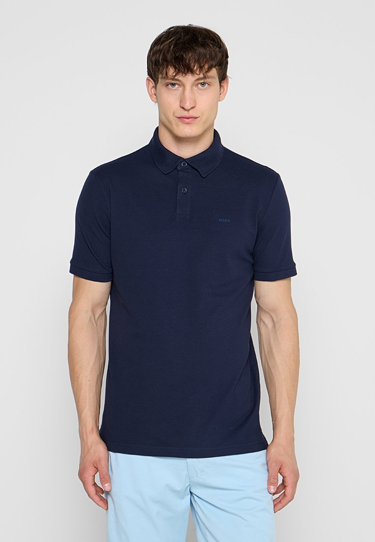 Junger Mann, der ein marineblaues kurzärmeliges Polohemd und hellblaue Hose trägt, steht mit dem Gesicht nach vorne vor einem einfarbigen Hintergrund.