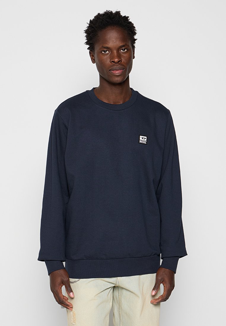 Junger Mann in einem navyblauen Diesel-Sweatshirt und hellbeigen, zerrissenen Jeans, der vor einem schlichten grauen Hintergrund steht.