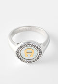 Silberner Ring mit rundem Gesicht, das ein goldfarbenes, stilisiertes „A“ zeigt, umgeben von kleinen klaren Edelsteinen auf weißem Hintergrund.