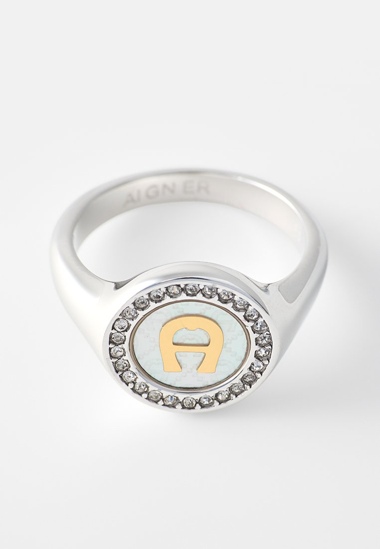 Silberner Ring mit rundem Gesicht, das ein goldfarbenes, stilisiertes „A“ zeigt, umgeben von kleinen klaren Edelsteinen auf weißem Hintergrund.