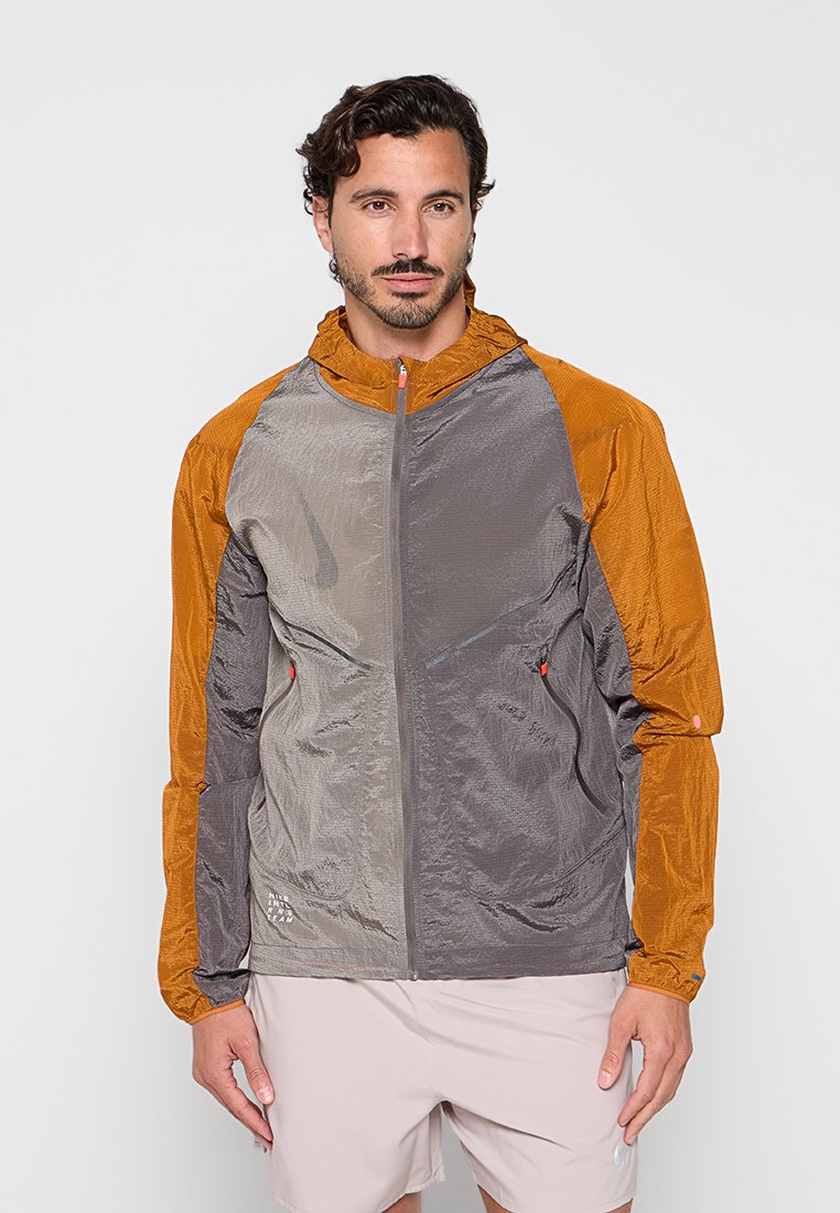 Mann trägt eine leichte Jacke mit orangen Ärmeln und grauem Körper, steht vor einem einfarbigen hellen Hintergrund.