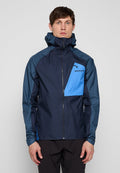 Mann trägt eine marineblaue und hellblaue Jacke mit Reißverschluss, hohem Kragen und Kapuze, steht vor einem schlichten grauen Hintergrund.