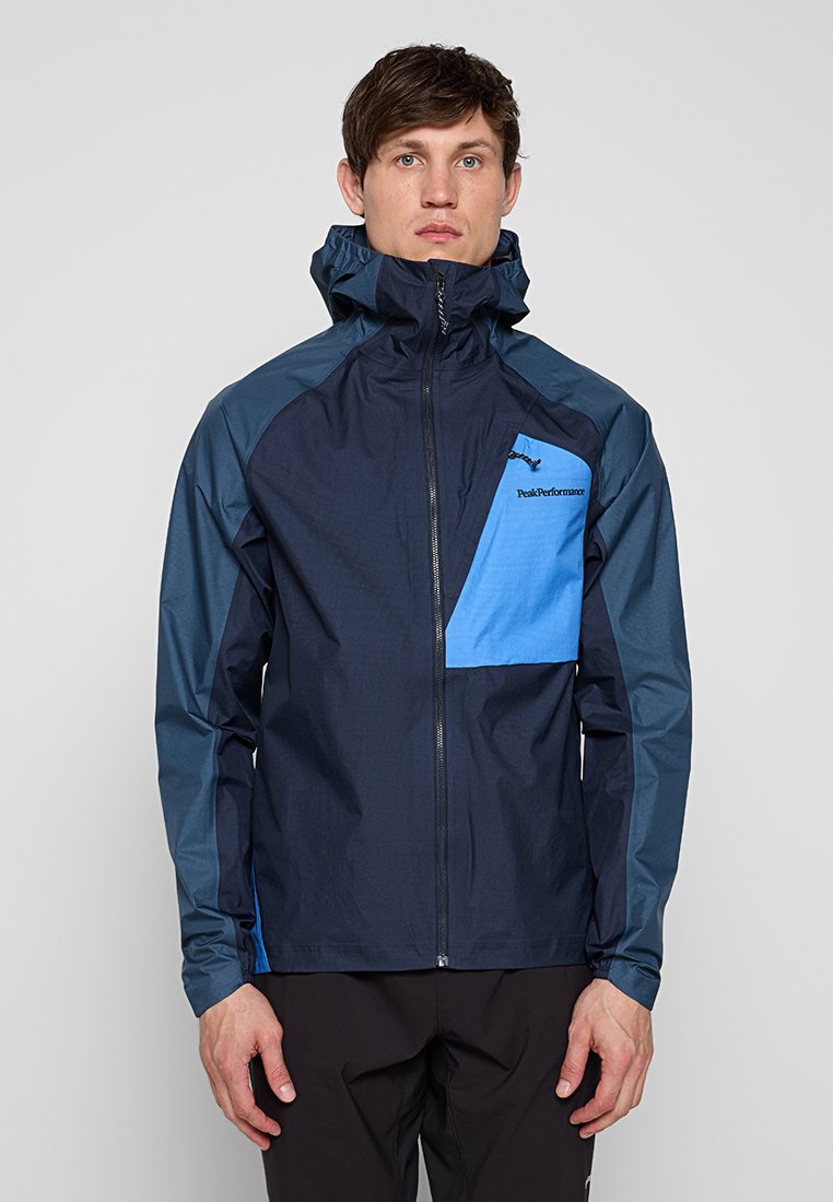 Mann trägt eine marineblaue und hellblaue Jacke mit Reißverschluss, hohem Kragen und Kapuze, steht vor einem schlichten grauen Hintergrund.