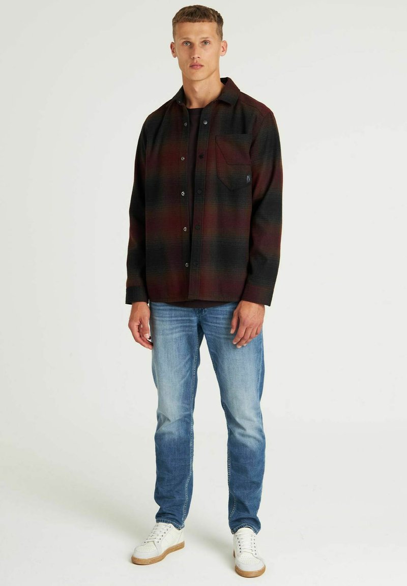 Kariertes Flanellhemd in Dunkelrot und Schwarz, mit einer Brusttasche und Knöpfen, kombiniert mit hellblauen Jeans und weißen Turnschuhen.