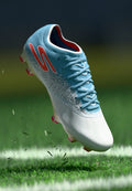 Blauer und weißer Fußballschuh mit strukturiertem Design, Schnürsenkeln und pinken Akzenten auf der Sohle. Der Schuh schwebt über dem Gras.