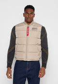 Mann trägt eine beige gesteppte Weste mit Reißverschluss und rotem Etikett über einem dunkelgrauen Pullover und blauen Jeans und steht vor einem schlichten hellen Hintergrund.