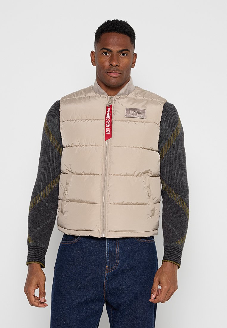 Mann trägt eine beige gesteppte Weste mit Reißverschluss und rotem Etikett über einem dunkelgrauen Pullover und blauen Jeans und steht vor einem schlichten hellen Hintergrund.
