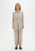 Hellbeige taillierter Blazer mit großen Knöpfen, kombiniert mit lockeren, hellbeigen Hosen mit elastischem Bündchen und seitlichen Taschen. Schwarze Schuhe.