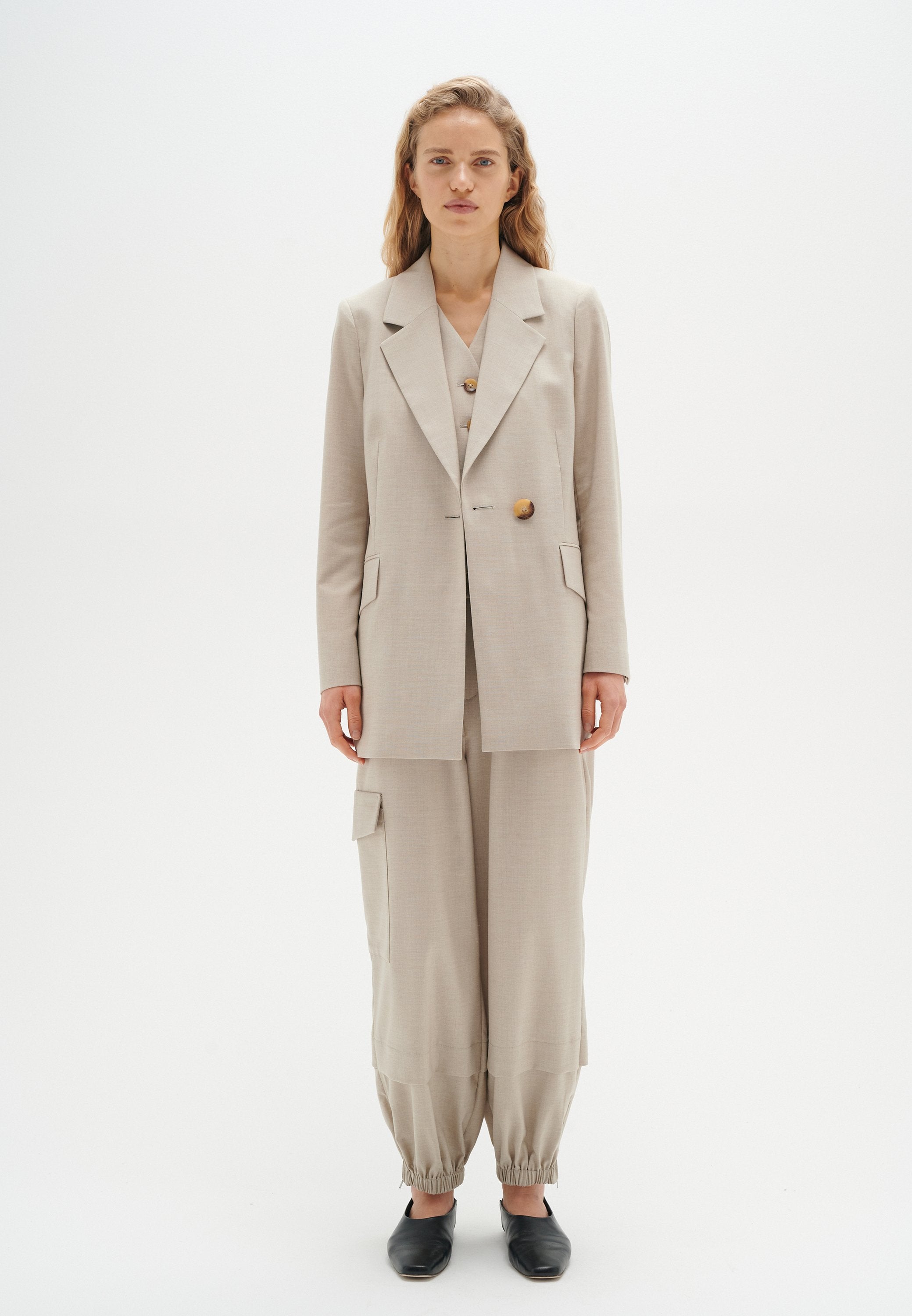 Hellbeige taillierter Blazer mit großen Knöpfen, kombiniert mit lockeren, hellbeigen Hosen mit elastischem Bündchen und seitlichen Taschen. Schwarze Schuhe.