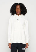 Junge Frau mit natürlichen lockigen Haaren, die einen weißen Nike-Hoodie und schwarze Hose trägt, steht vor einem einfarbigen hellen Hintergrund.