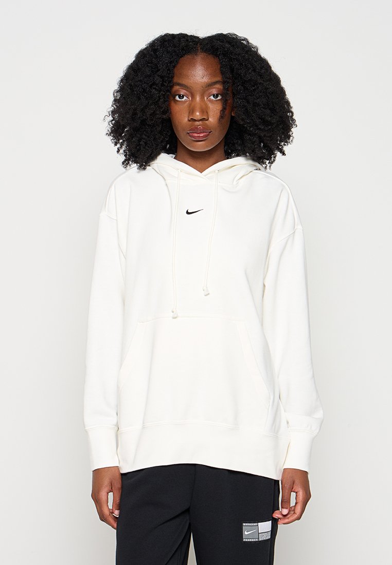 Junge Frau mit natürlichen lockigen Haaren, die einen weißen Nike-Hoodie und schwarze Hose trägt, steht vor einem einfarbigen hellen Hintergrund.