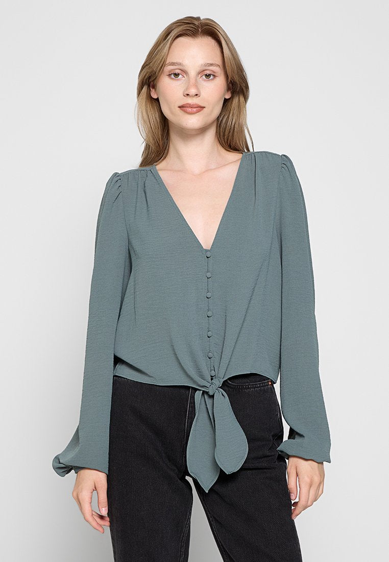 Tiefgrüne Langarm-Bluse mit tiefem V-Ausschnitt, Knopfverschluss vorne und gebundenem Knoten-Detail an der Taille. Weicher, leichter Stoff mit leichter Struktur.