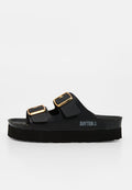 Schwarze Slip-On-Sandale mit zwei verstellbaren goldenen Schnallen und dicker Sohle, mit dem Markennamen Bayton und einem Ankerlogo an der Seite.