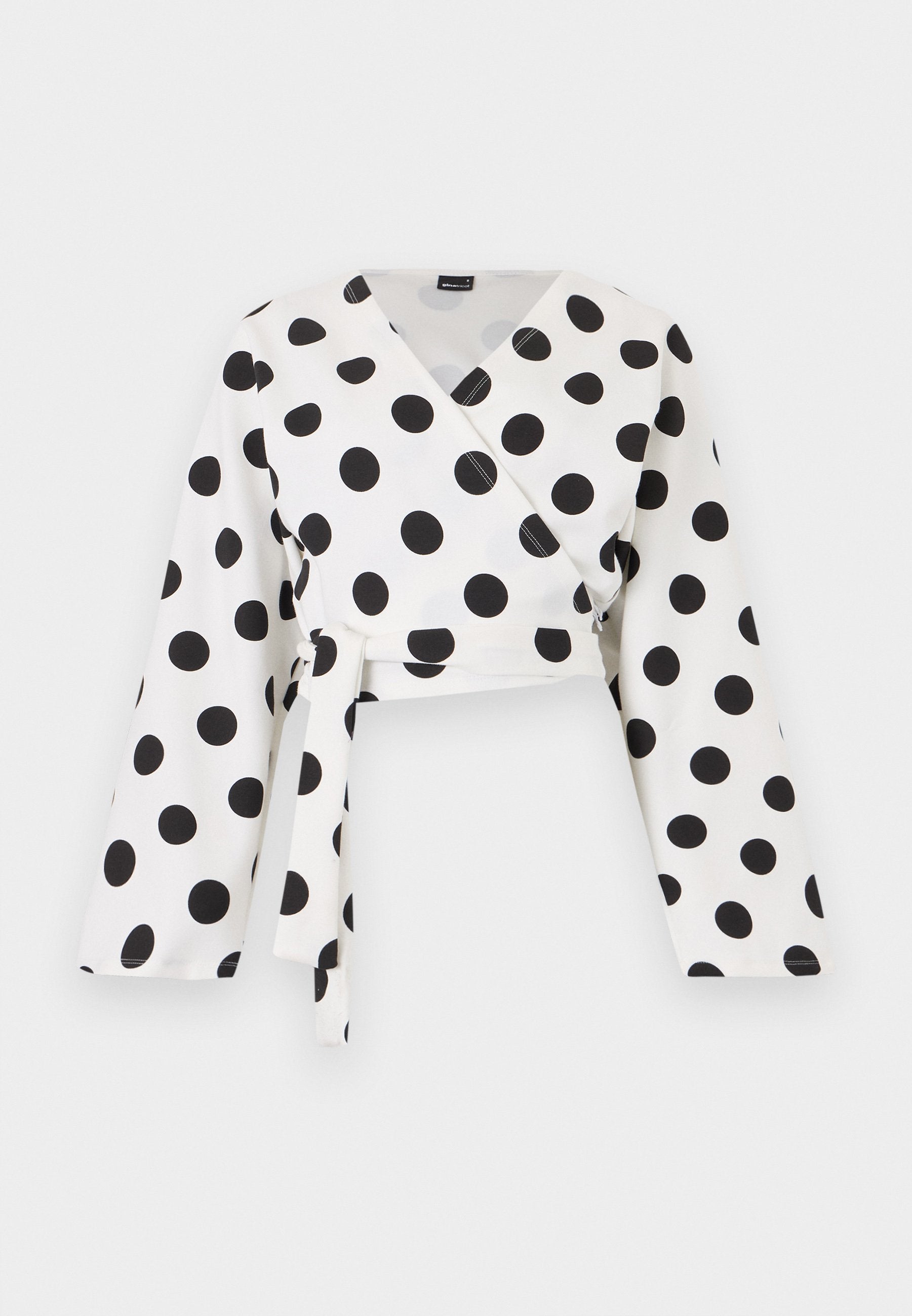 Weiße Langarm-Wickelbluse mit großen schwarzen Polka-Dots und einem vorn gebundenen Gürtel, dargestellt auf einfachem weißem Hintergrund.