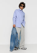 Mann mit lockigem Haar und Sonnenbrille, der ein hellblaues, kurzärmeliges Hemd, graue weit geschnittene Hosen und silberne Sneakers trägt, während er eine blaue Jeansjacke hält.