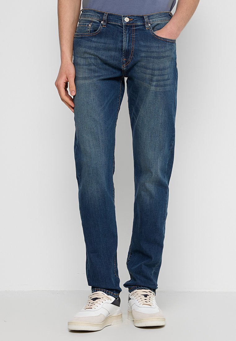 Blaue Jeans aus Denim mit geradem Schnitt, sichtbaren Nähten, einem Knopfverschluss vorne und fünf Taschen, kombiniert mit weißen Turnschuhen.