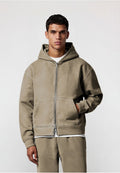 Junger Mann trägt einen beigen Zip-Hoody und passende Hose, mit einer Hand in der Tasche, steht vor einem schlichten hellen Hintergrund.