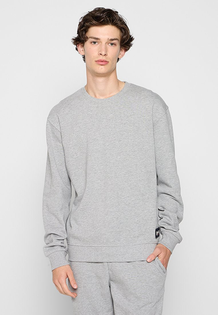 Junger Mann mit lockigem Haar trägt einen hellgrauen Sweatshirt und passende Jogginghose, steht vor einem schlichten weißen Hintergrund.