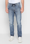 Mann in verwaschenen blauen Denim-Jogginghosen mit Kordelzug und weißen Turnschuhen, der vor einem einfarbigen hellen Hintergrund steht.