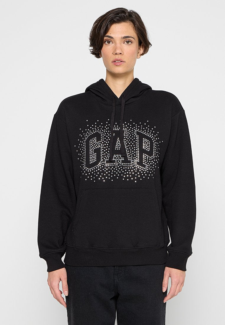 Person trägt schwarzen Hoodie mit silbernen, mit Nieten besetzten "GAP"-Logo, steht vor einfarbigem hellgrauem Hintergrund.