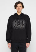 Person trägt schwarzen Hoodie mit silbernen, mit Nieten besetzten "GAP"-Logo, steht vor einfarbigem hellgrauem Hintergrund.