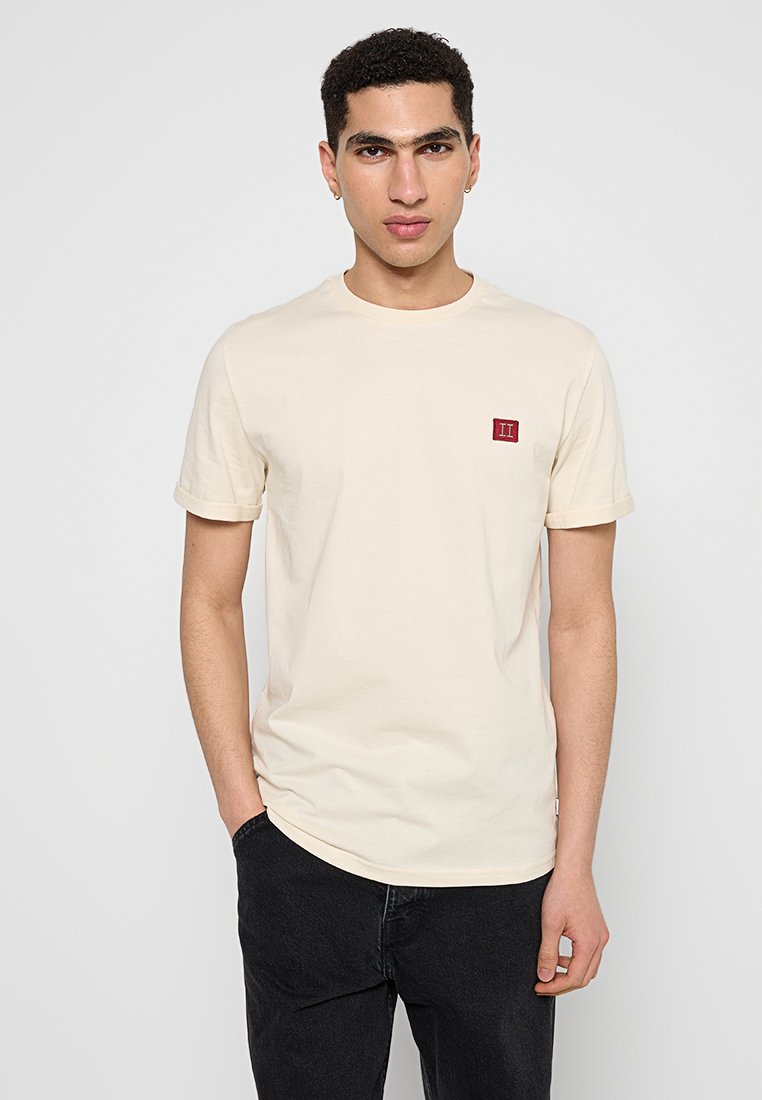 Junger Mann mit kurzen lockigen Haaren, der ein beiges Kurzarm-T-Shirt mit einem kleinen roten Patch und schwarze Hosen trägt, steht mit einer Hand in der Tasche.