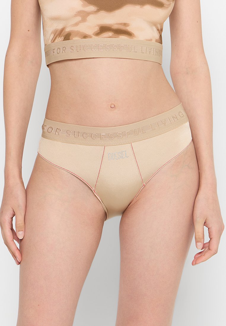 Beige Unterwäsche mit einer glatten, glänzenden Textur, die einen breiten Bund aufweist, der mit "FÜR EINEN ERFOLGREICHEN LEBENSSTIL" beschriftet ist und mit kontrastierendem roten Nähten versehen ist.