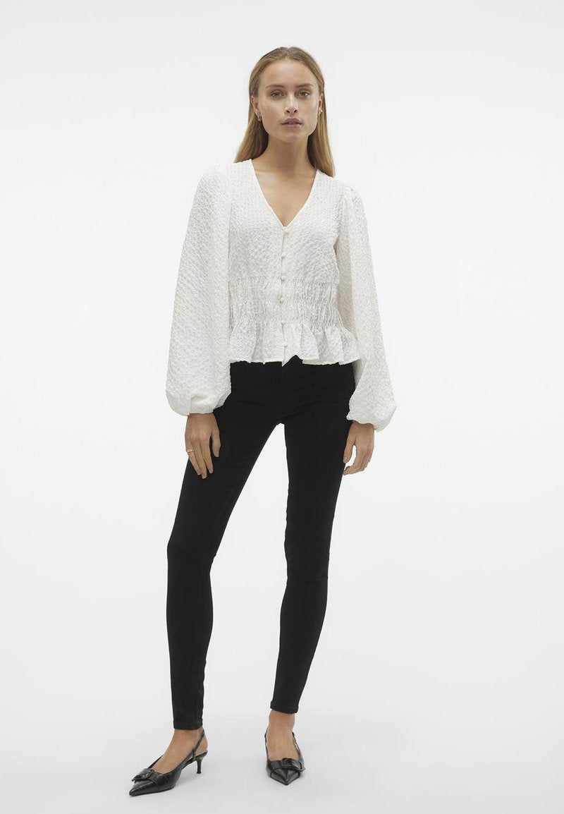 Weiße Peplum-Bluse aus strukturiertem Stoff, mit V-Ausschnitt und Knopfverschluss; kombiniert mit schwarzen, engen Hosen und schwarzen, spitz zulaufenden Schuhen.