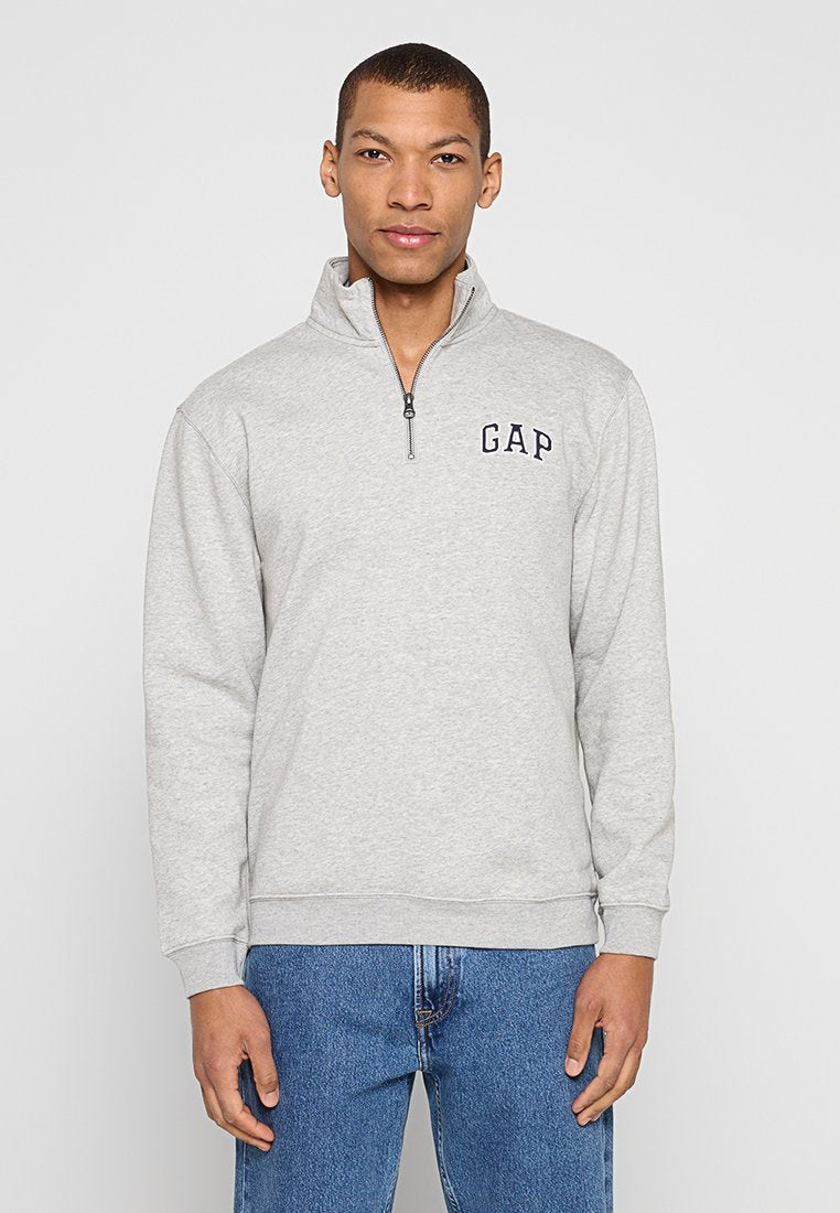 Grauer Pullover mit einem halbhohen Reißverschlusskragen. Verfügt über ein marineblaues "GAP"-Logo auf der Brust und gerippte Bündchen. Wird mit blauen Jeans getragen.