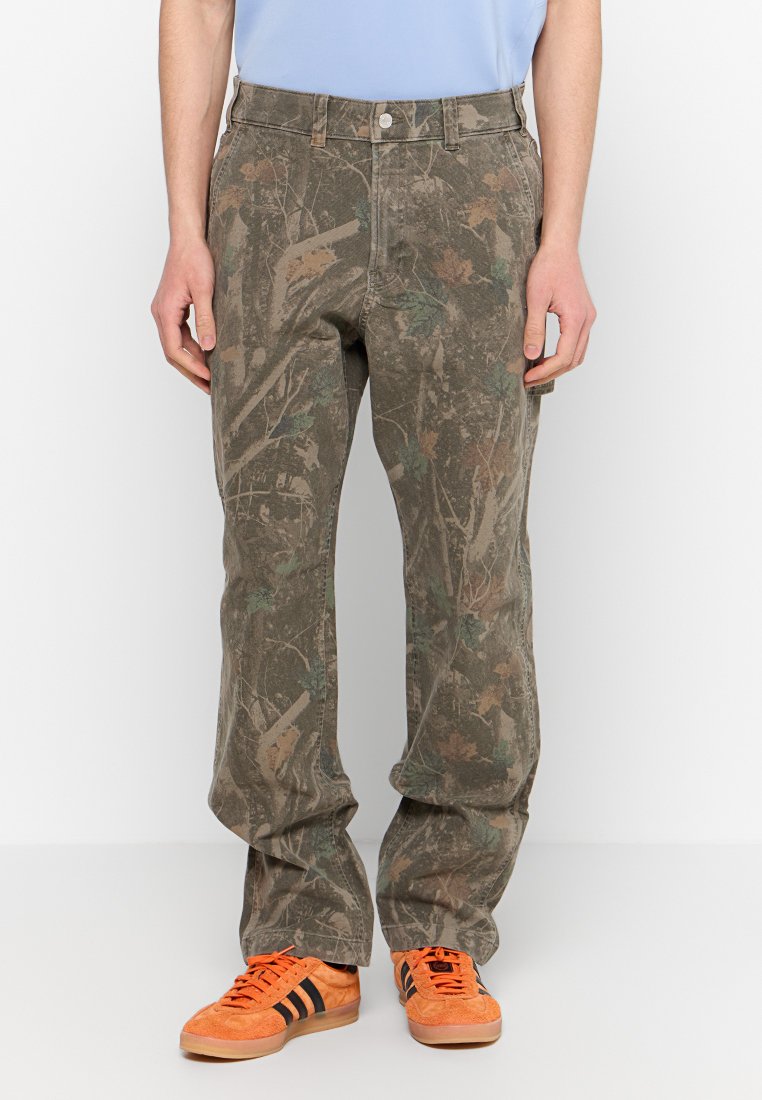 Herren-Camouflage-Hosen in erdigen Tönen mit weiter Passform. Besitzt Taschen und einen Knopfverschluss. Kombiniert mit orangefarbenen Sneakers für einen Kontrast.