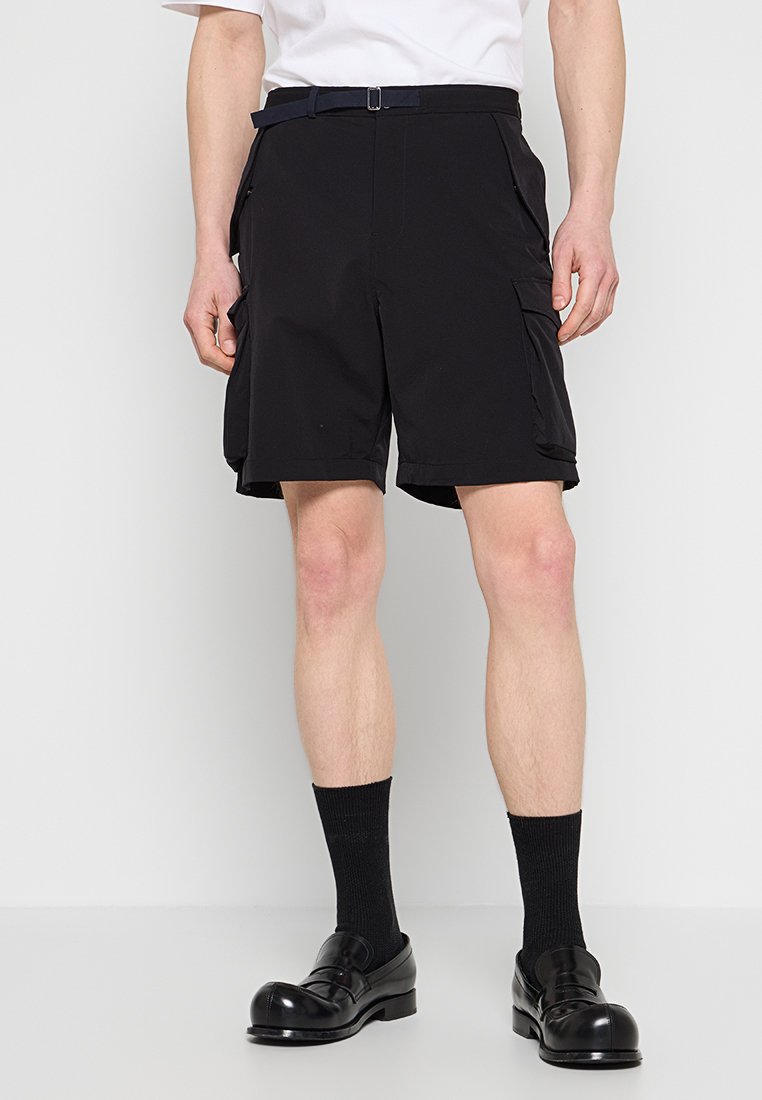 Schwarze Cargo-Shorts mit Gürtel, seitlichen Taschen und lässiger Passform, aus leichtem Material; kombiniert mit schwarzen Socken und Schuhen.