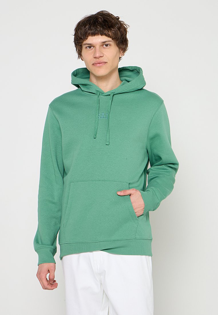 Junger Mann mit kurzen lockigen Haaren, der einen grünen Hoodie mit Fronttasche und weiße Hose trägt, vor einem schlichten hellen Hintergrund stehend.